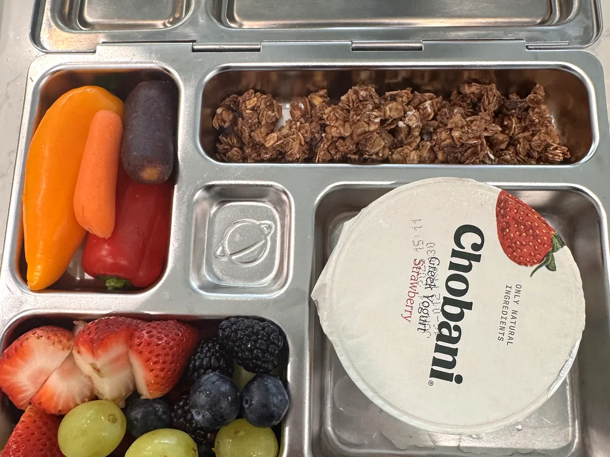 A steel bento box with a yogurt cup, granola, berries and grapes, carrots and peppers